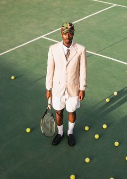 Man in blazer and shorts holding tennis racket on outdoor court surrounded by balls.