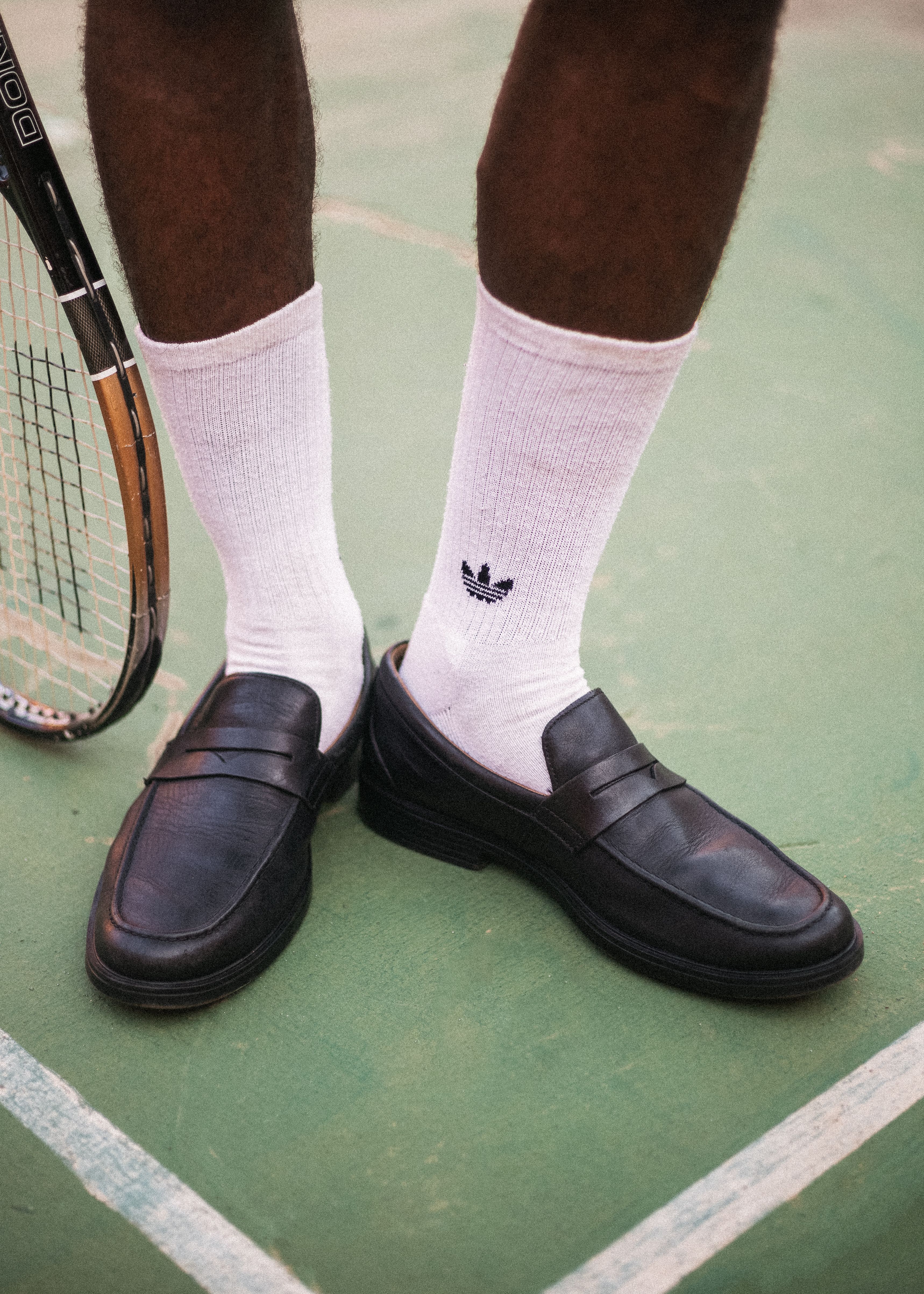 A person wearing black loafers and white socks stands on a tennis court, holding a racket.