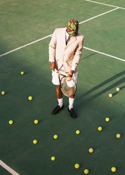A fashionable man in a suit stands on a tennis court surrounded by tennis balls.