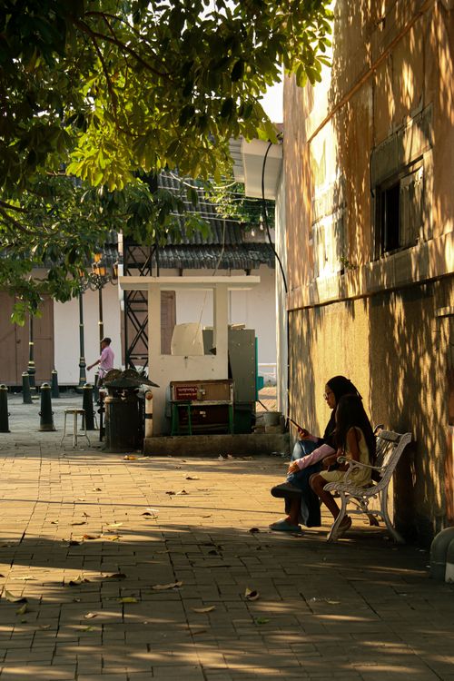 Free Two people relax on a bench in a sunlit urban area with trees and shadows. Stock Photo