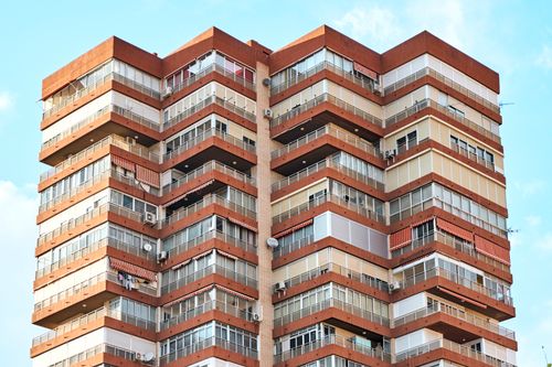 Free Close-up of a modern multi-story residential building with red and beige facade against a clear blue sky. Stock Photo