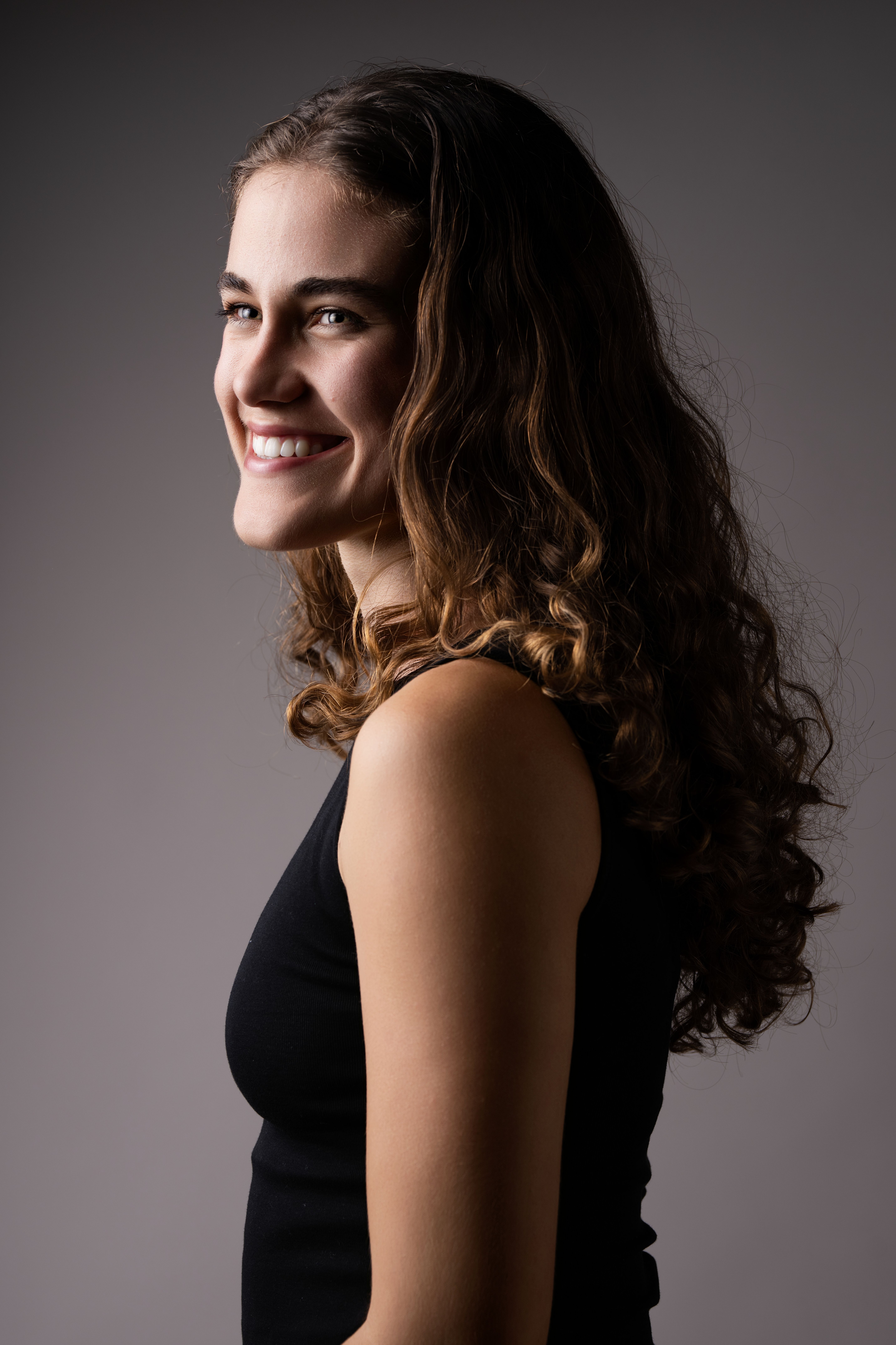 Side profile of a smiling woman in a black dress with curly hair, shot in a studio setting.