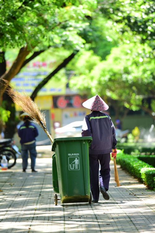 Free Street cleaner pushes a bin while wearing a traditional conical hat in a sunny park. Stock Photo