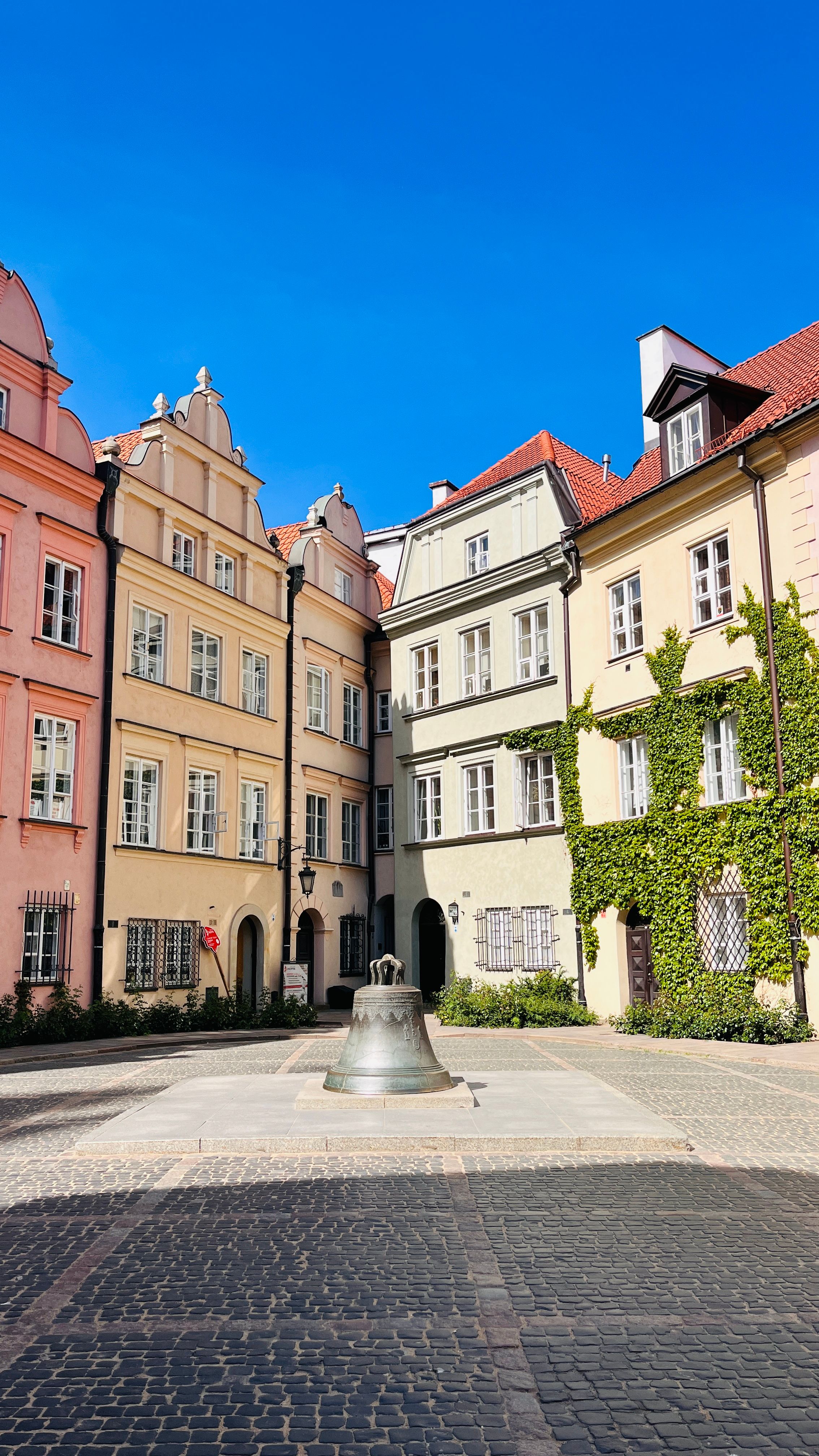 Scenic view of a European courtyard with colorful historic buildings, blue sky, and a bell in the center.