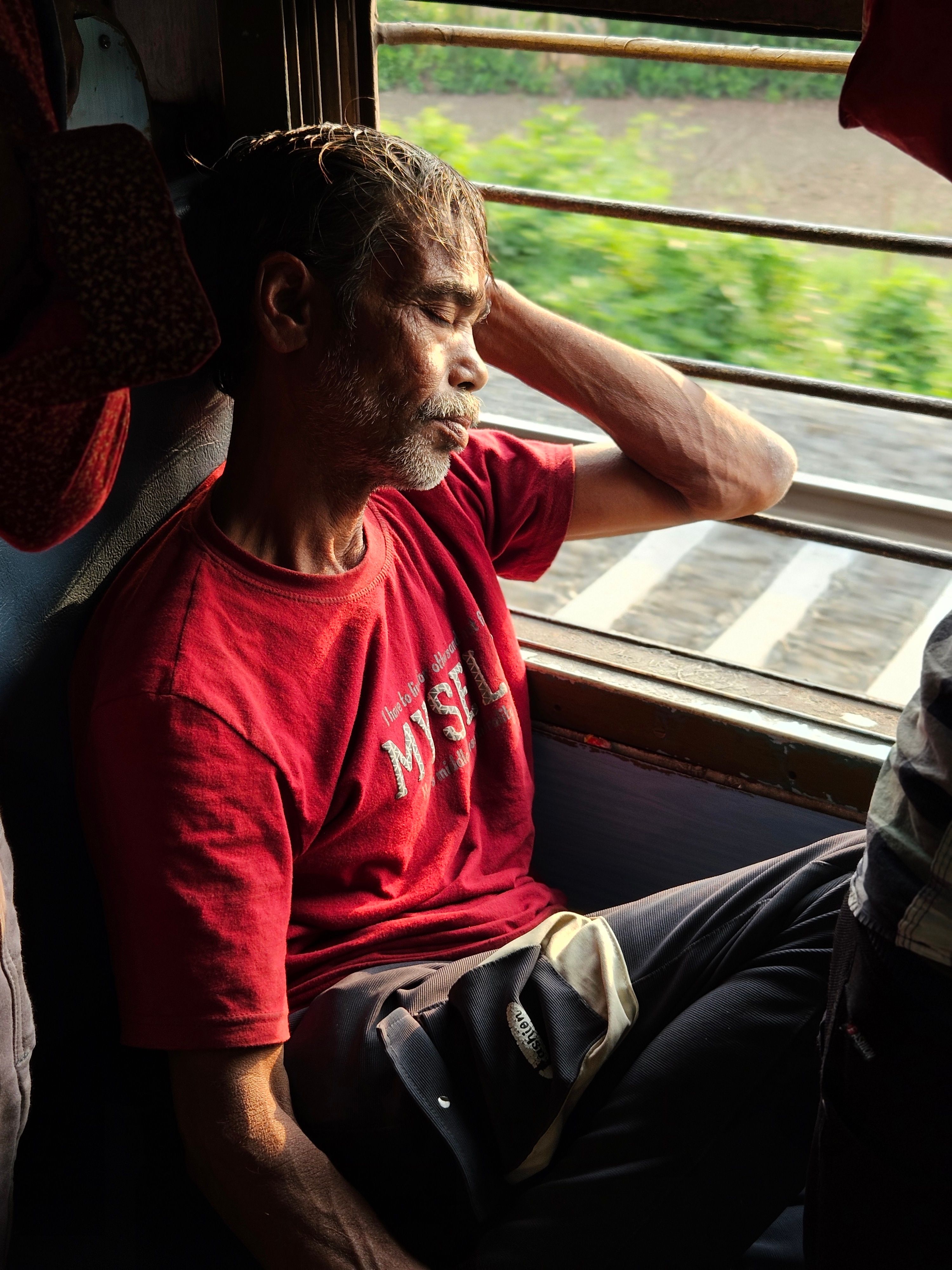 Man in red shirt resting by train window, immersed in thought during the daytime journey.