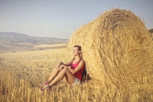 Woman in Red Tank Top Sitting on Brown Hay Field Leaning on Hay Roll