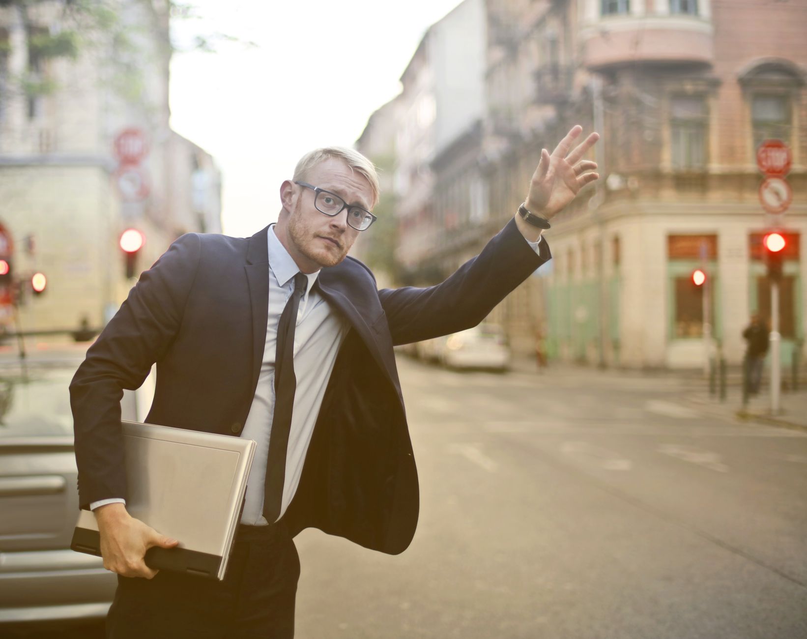 A confident businessman in a suit hails a taxi on a bustling city street, holding a laptop.