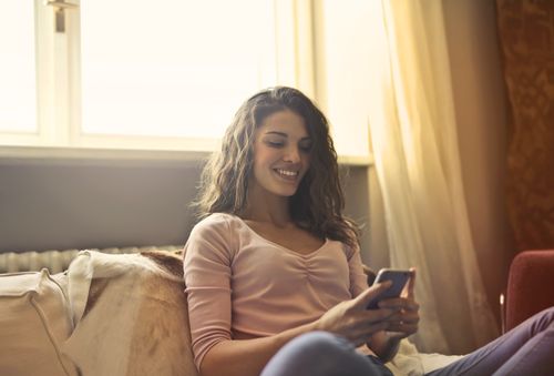 Free Smiling woman sits comfortably indoors, enjoying leisure time with her smartphone near a window. Stock Photo