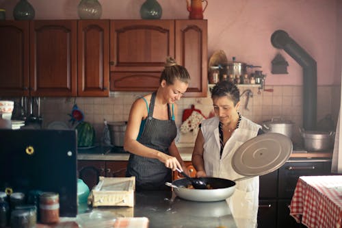 Daughter and senior mother standing at table in kitchen and stirring dish in frying pan while preparing food for dinner