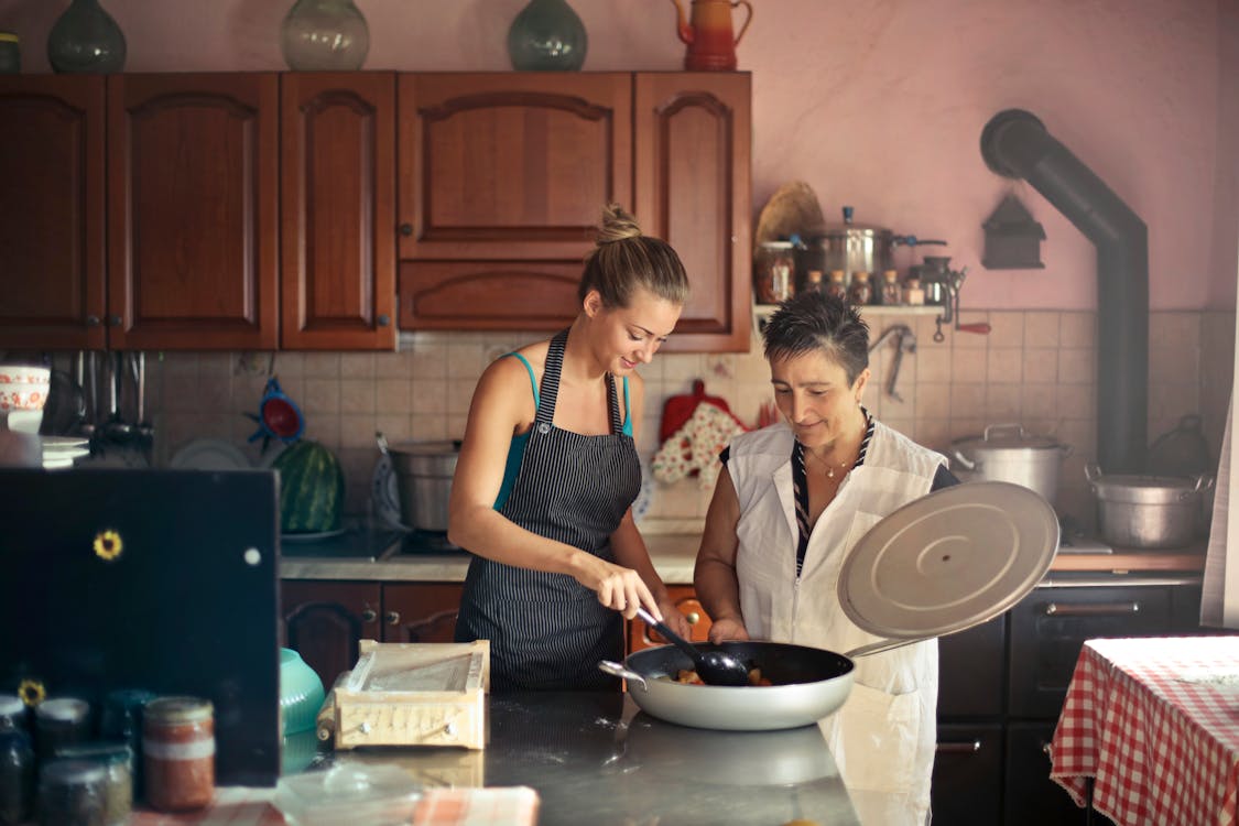 Free Daughter and senior mother standing at table in kitchen and stirring dish in frying pan while preparing food for dinner Stock Photo