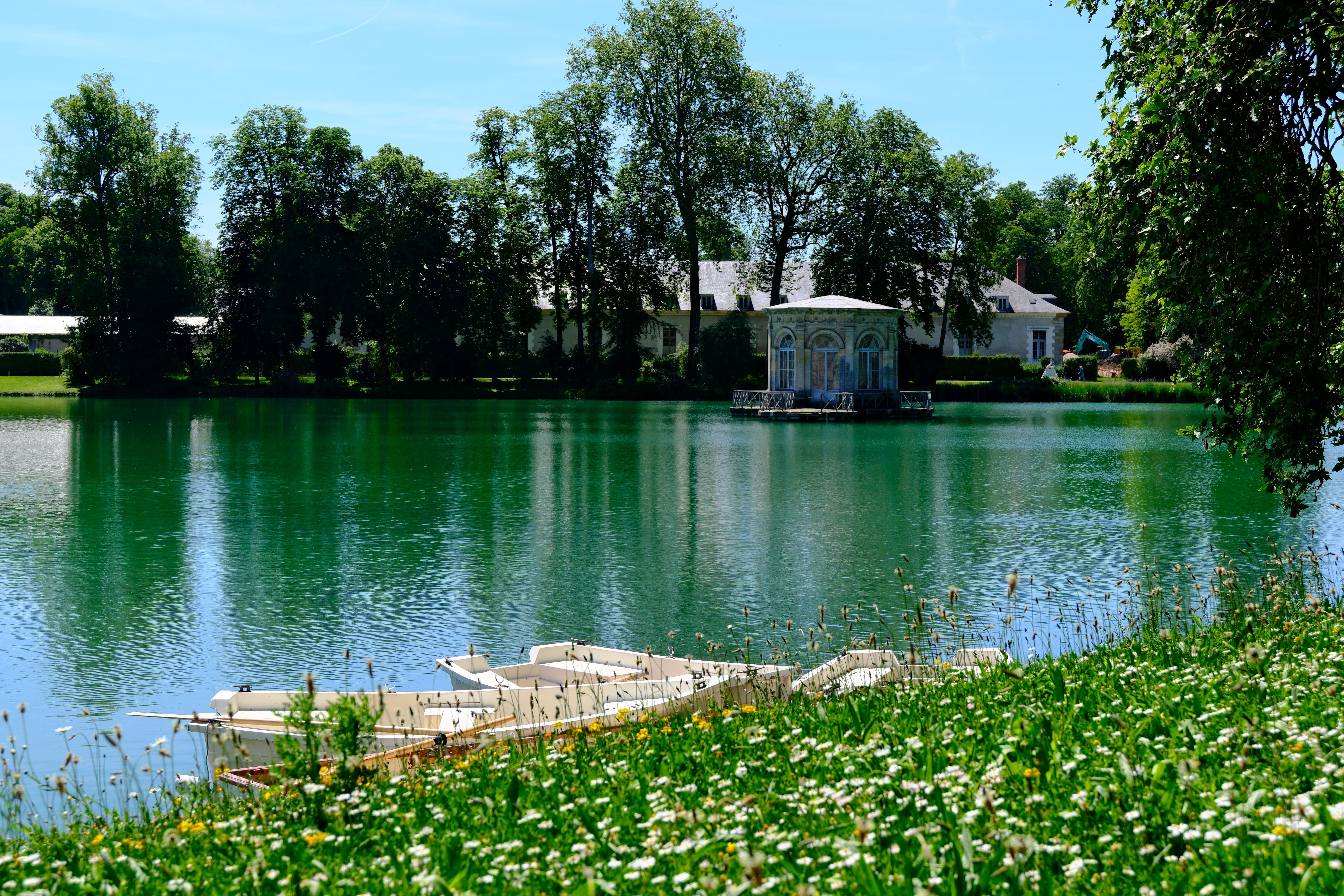 Sunlit lake in Fontainebleau, France, with boats, a classical pavilion, tree-lined architecture and distant visitors
