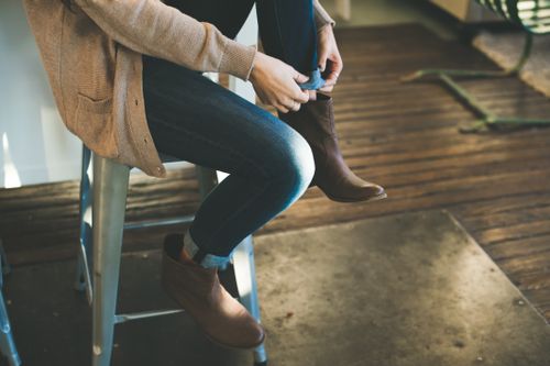Free A woman sitting on a stool indoors, putting on brown leather boots with casual attire. Stock Photo