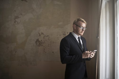 Free Side view of young distracted male entrepreneur in classy suit standing near window in shabby house and reading messages about eviction Stock Photo