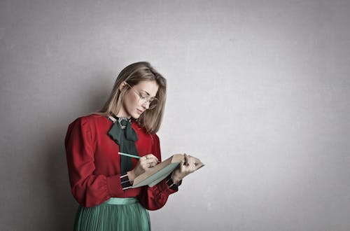 Free Pensive attentive woman in glasses and elegant vintage outfit focusing and taking notes with pencil in book while standing against gray wall Stock Photo