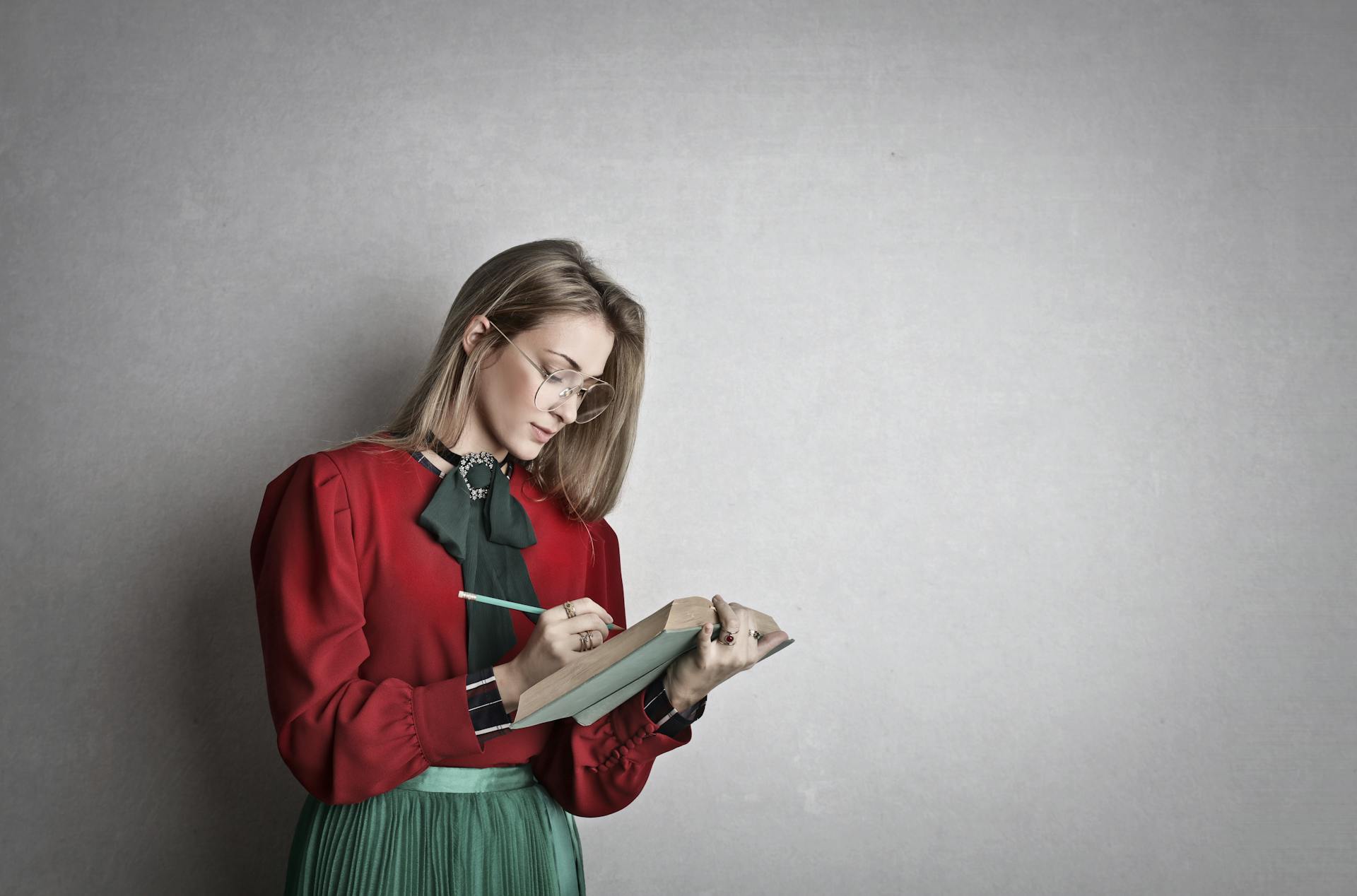 Pensive attentive woman in glasses and elegant vintage outfit focusing and taking notes with pencil in book while standing against gray wall