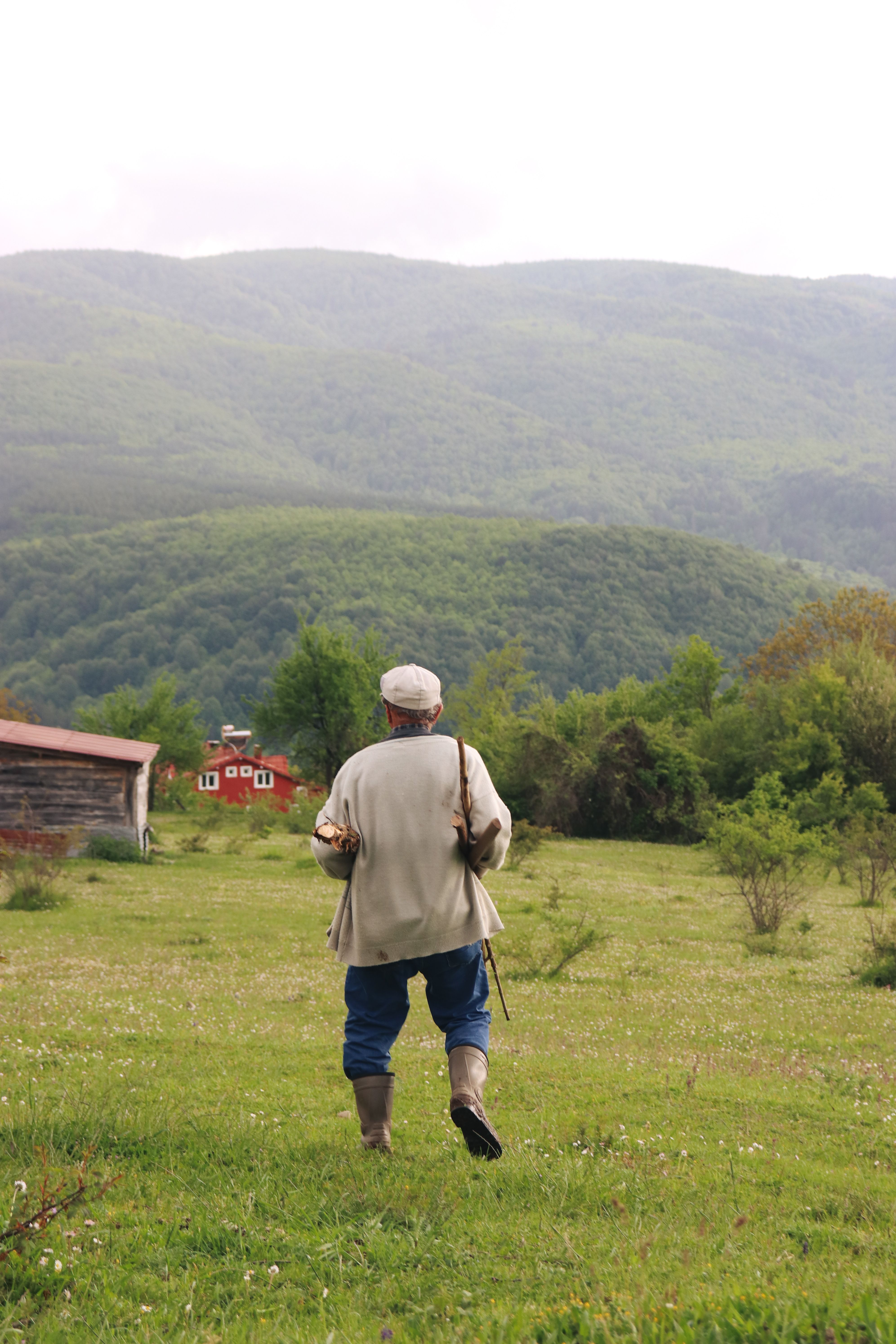An elderly farmer walks through a lush green field towards a red barn with mountainous background.