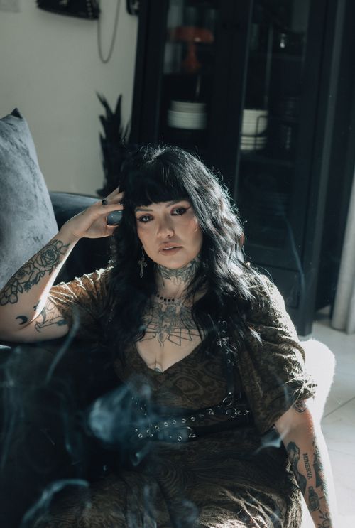 Free A woman with tattoos sits indoors, looking contemplatively into the distance, surrounded by soft natural lighting. Stock Photo