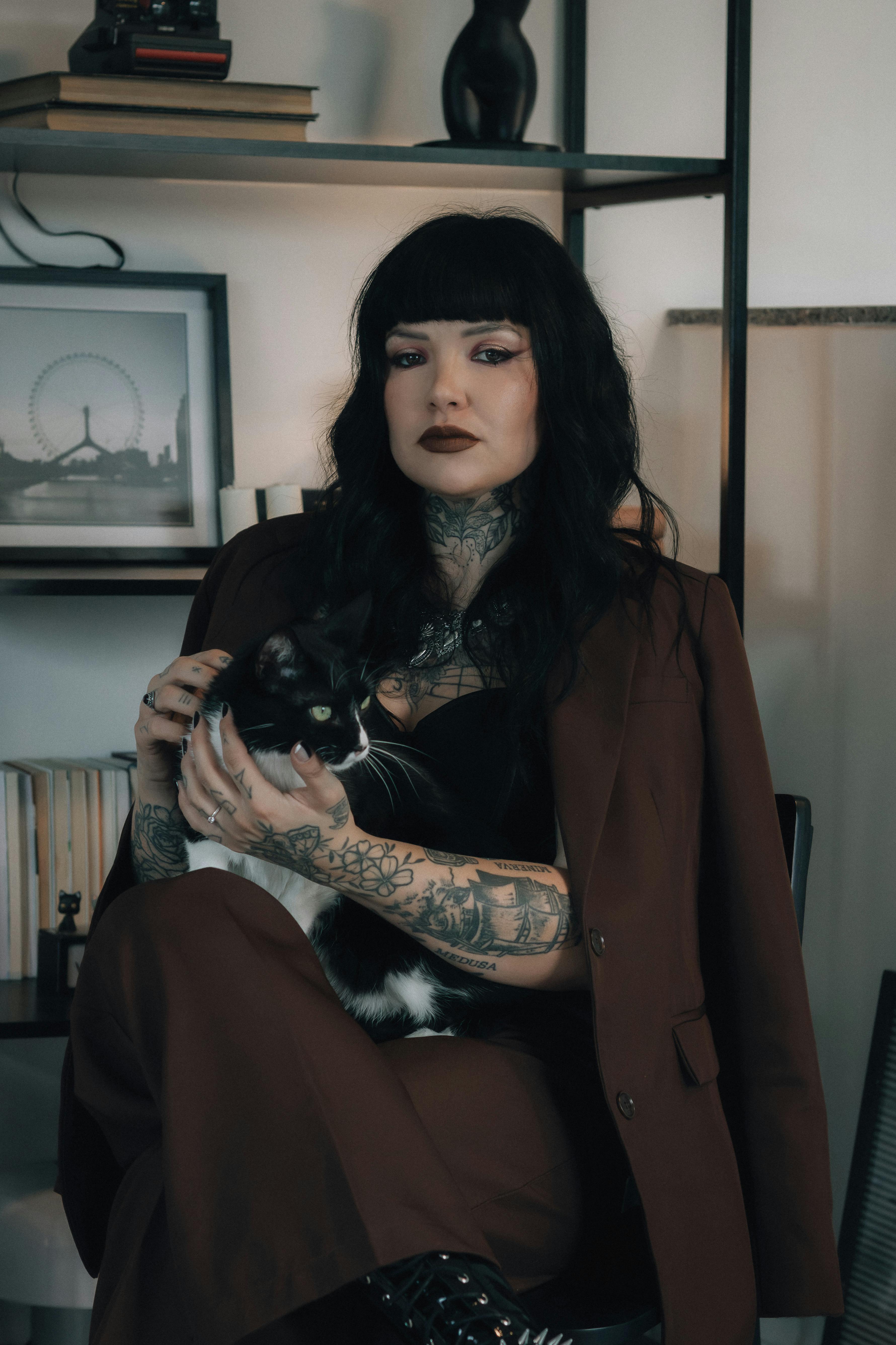 A tattooed woman in a brown suit holding a black and white cat in a modern indoor setting.