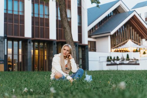 Free A young woman sitting on grass in front of a modern building with illuminated lights in Minsk, Belarus. Stock Photo