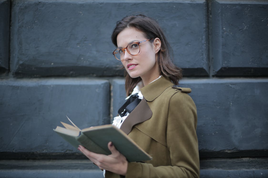 Free From below confident young female in glasses and elegant vintage clothes looking away and contemplating while reading rook on street near black wall of aged building in city Stock Photo