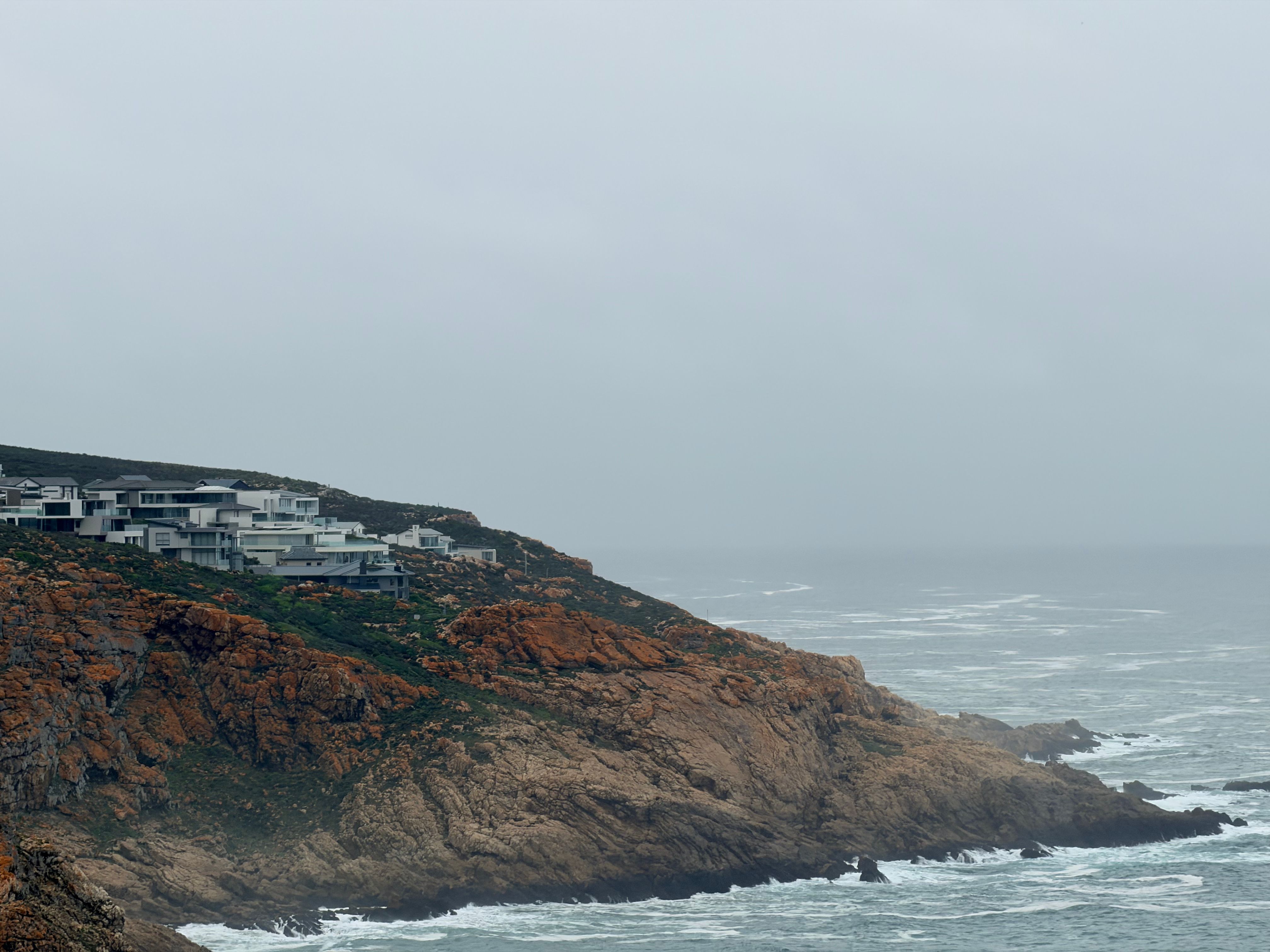 Scenic view of modern homes perched on rocky cliffs overlooking a misty ocean in George, South Africa.