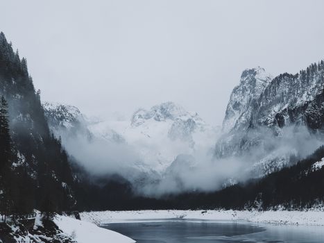 Majestic snowy mountains with misty clouds and serene icy lake in winter landscape.