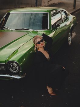 Stylish woman in black poses by a classic green car on a street in Brazil, exuding retro charm.
