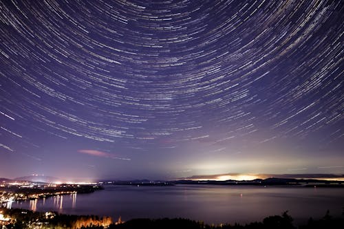 Free Amazing view of glowing stars in long exposure above calm sea and city located on shore Stock Photo