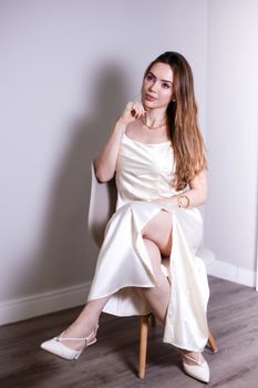 A woman in an elegant white satin dress sits gracefully on a chair indoors.