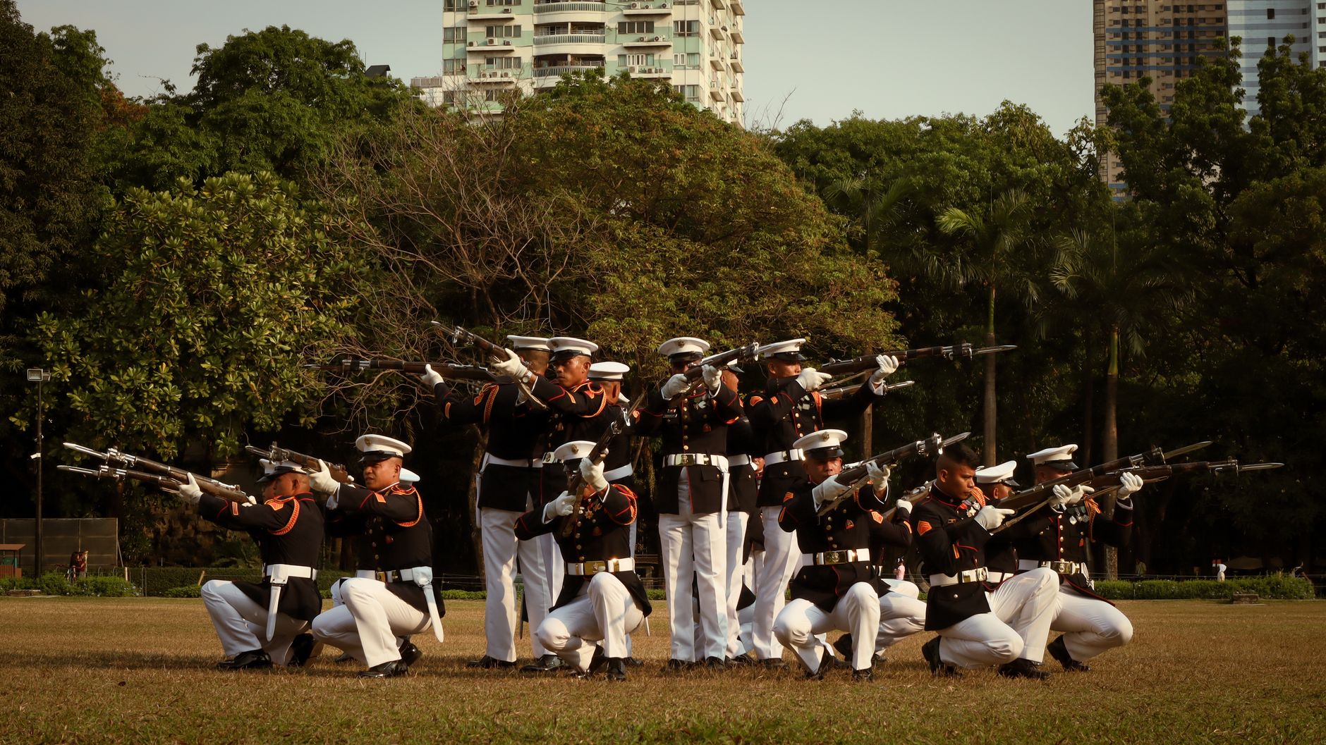 Uniformed soldiers performing a drill in a park with urban backdrop.