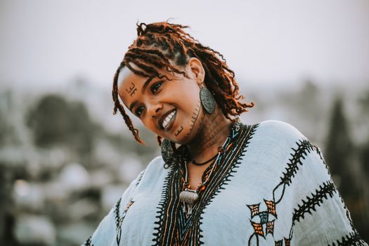 A smiling woman with traditional tattoos and Ethiopian attire enjoying a sunny day outdoors.