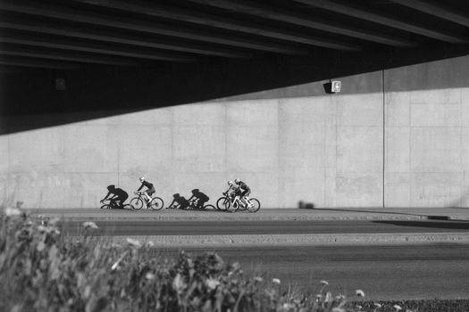 Black and white photo of cyclists racing under a bridge, casting shadows on a concrete wall.
