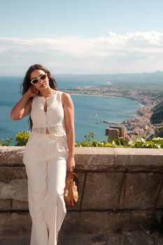 Fashionable woman in white attire poses against a stunning coastal backdrop on a sunny day.