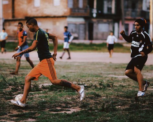 Free Energetic young adults playing a lively game of soccer in an urban setting. Grass and dirt field with a brick building backdrop. Stock Photo