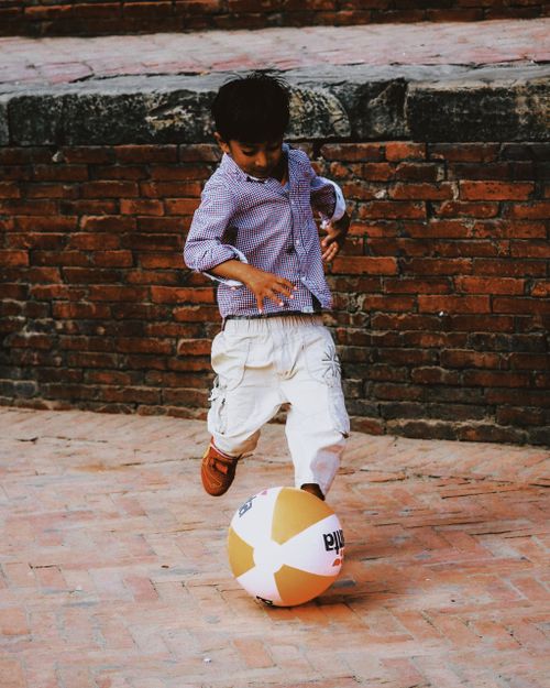 Free A boy in casual attire kicking a large ball on a brick sidewalk. Stock Photo