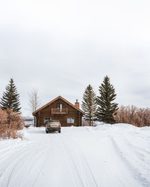 Idyllic winter scene of a cabin with parked car under a snowy landscape and pine trees.