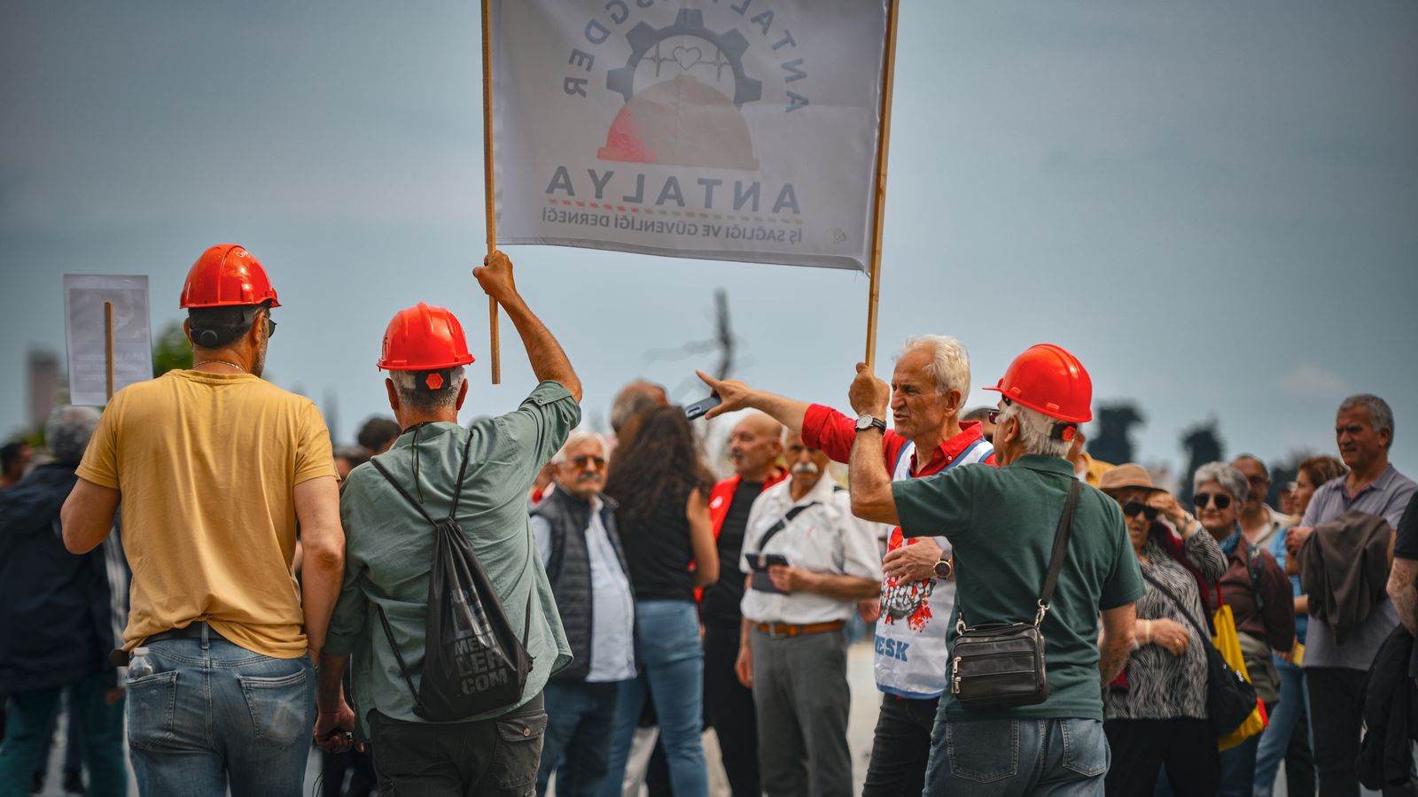 A group of people participating in a protest in Antalya, Türkiye, wearing red hard hats and holding signs.