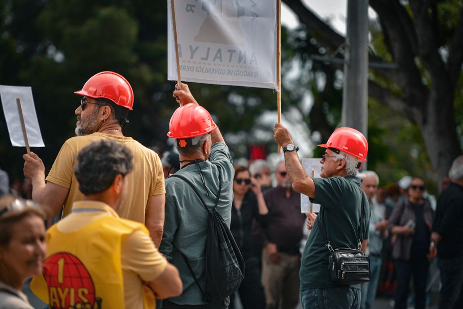 Protesters wearing red hard hats and holding signs gather in Antalya, Türkiye, highlighting activism.