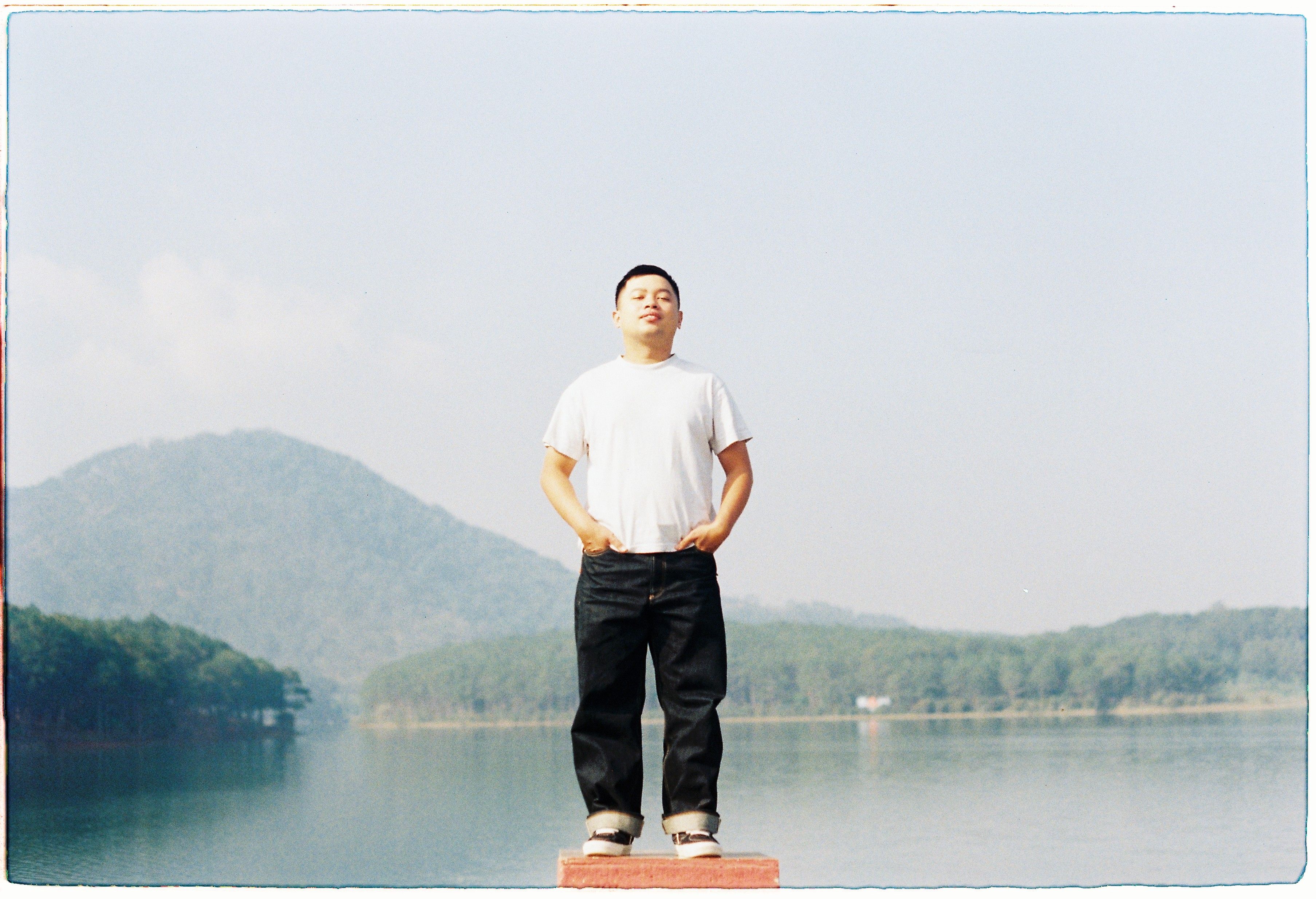 A man stands calmly by a serene lake in Ho Chi Minh City with mountains in the background.