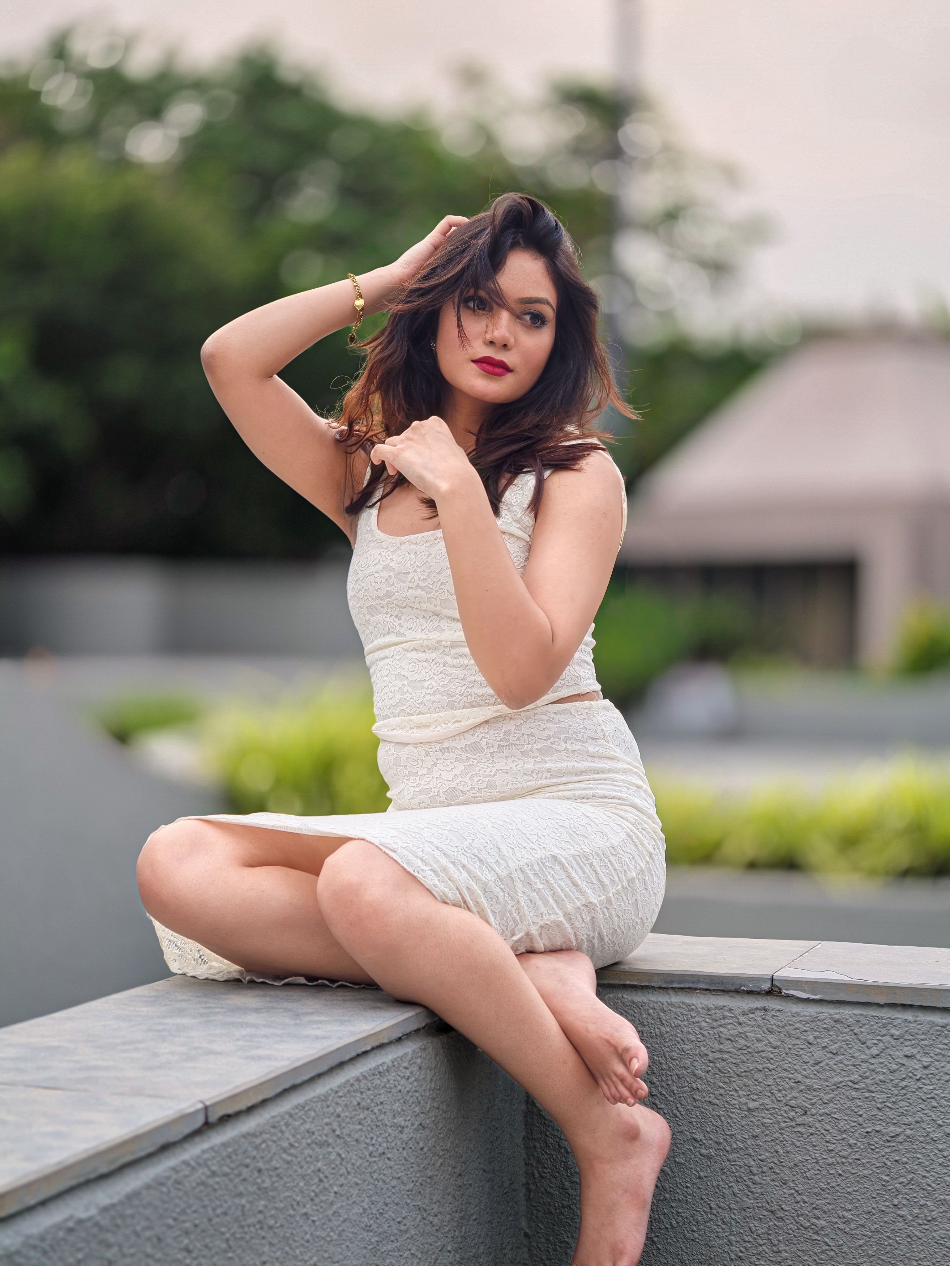Elegant woman in a white dress poses gracefully on a ledge outdoors, exuding confidence.