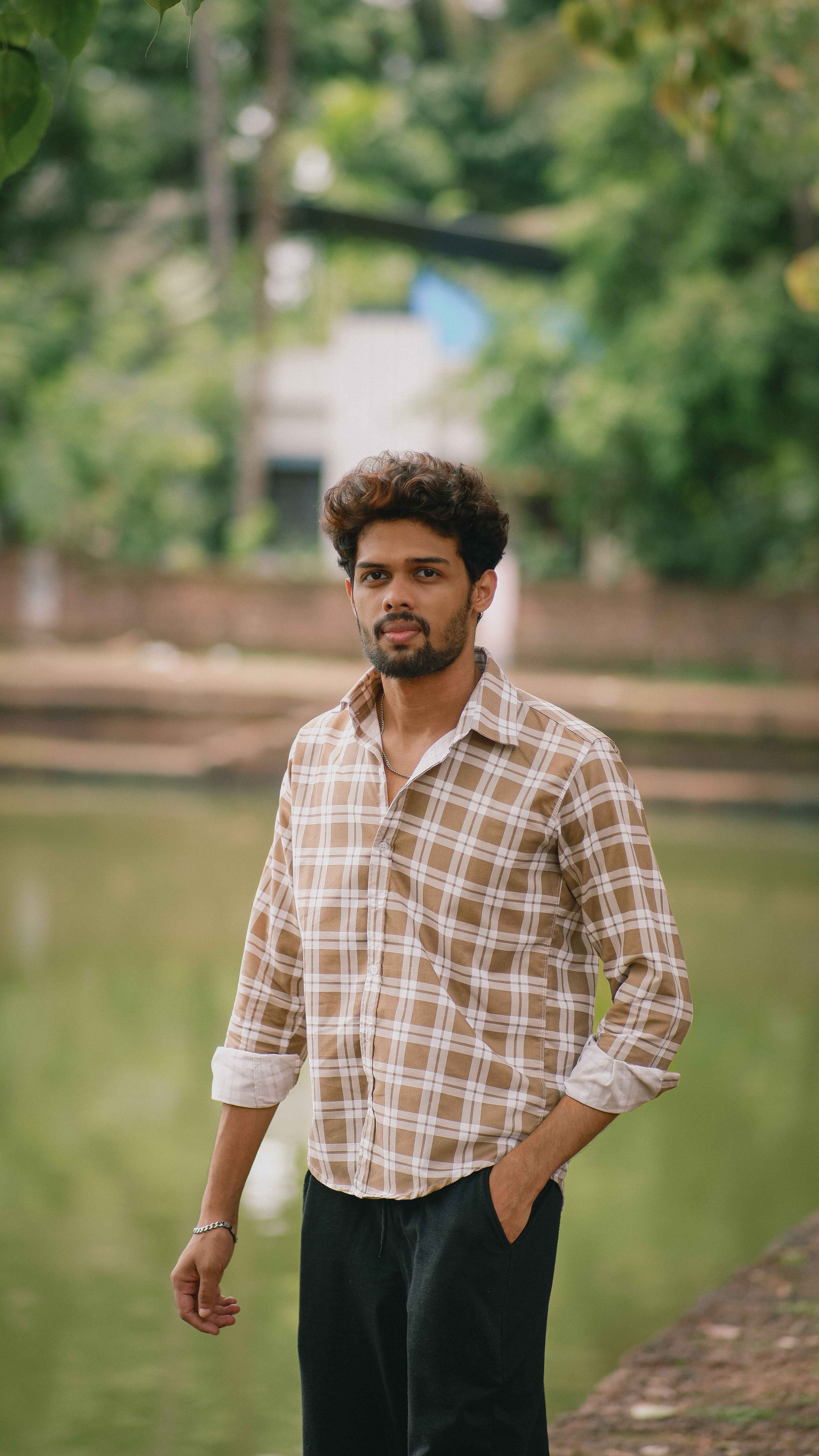 A man in plaid shirt stands by a river, surrounded by lush greenery.