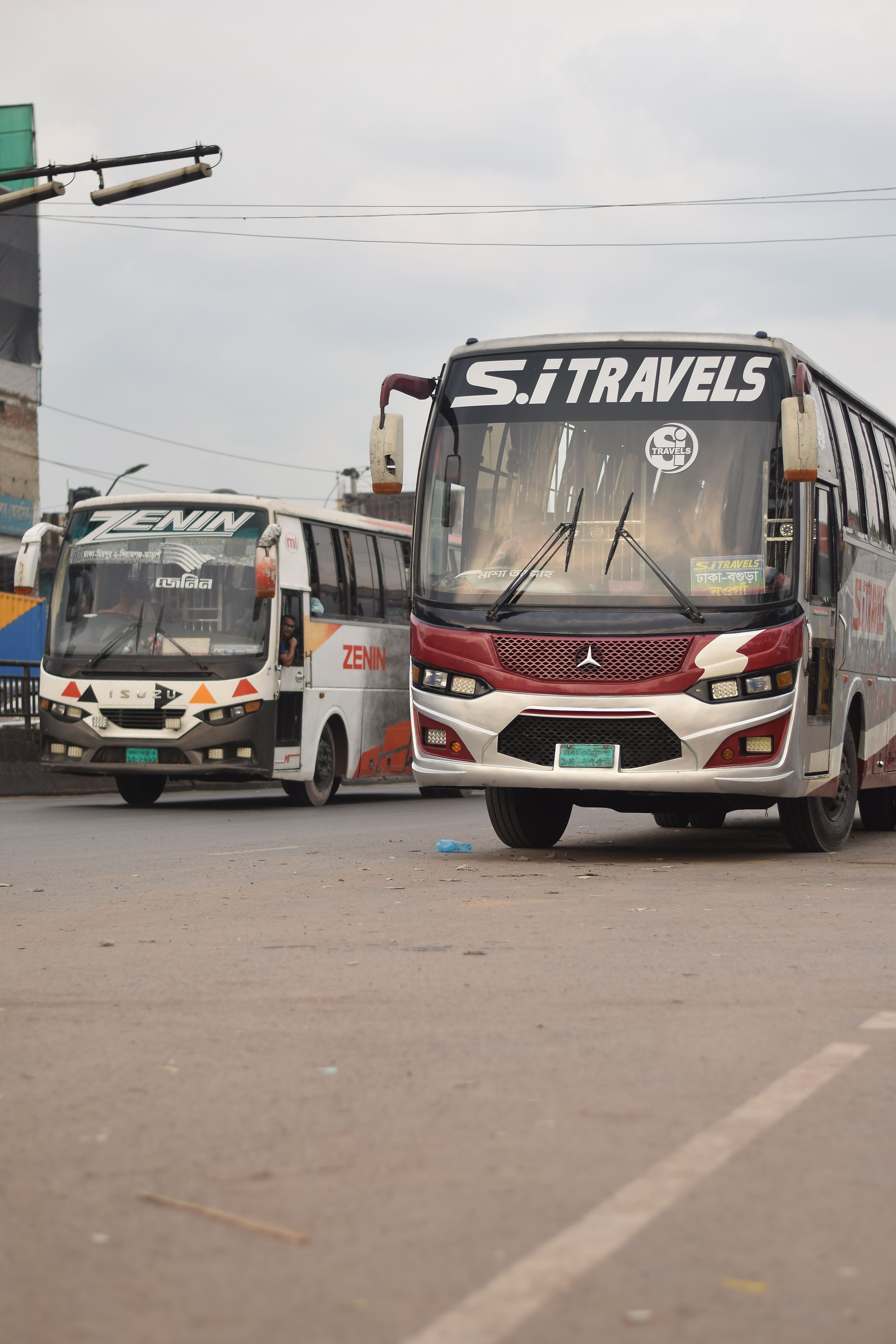 Two public buses parked on a city street in Dhaka, Bangladesh, with visible brand names.