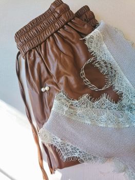 Stylish flat lay of brown leather pants, lace top, pearl earring, and woven bracelet.