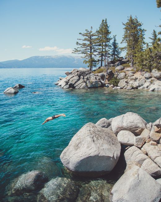 Distant unrecognizable person jumping into turquoise water from stone shore of lake located in mountainous area with forest