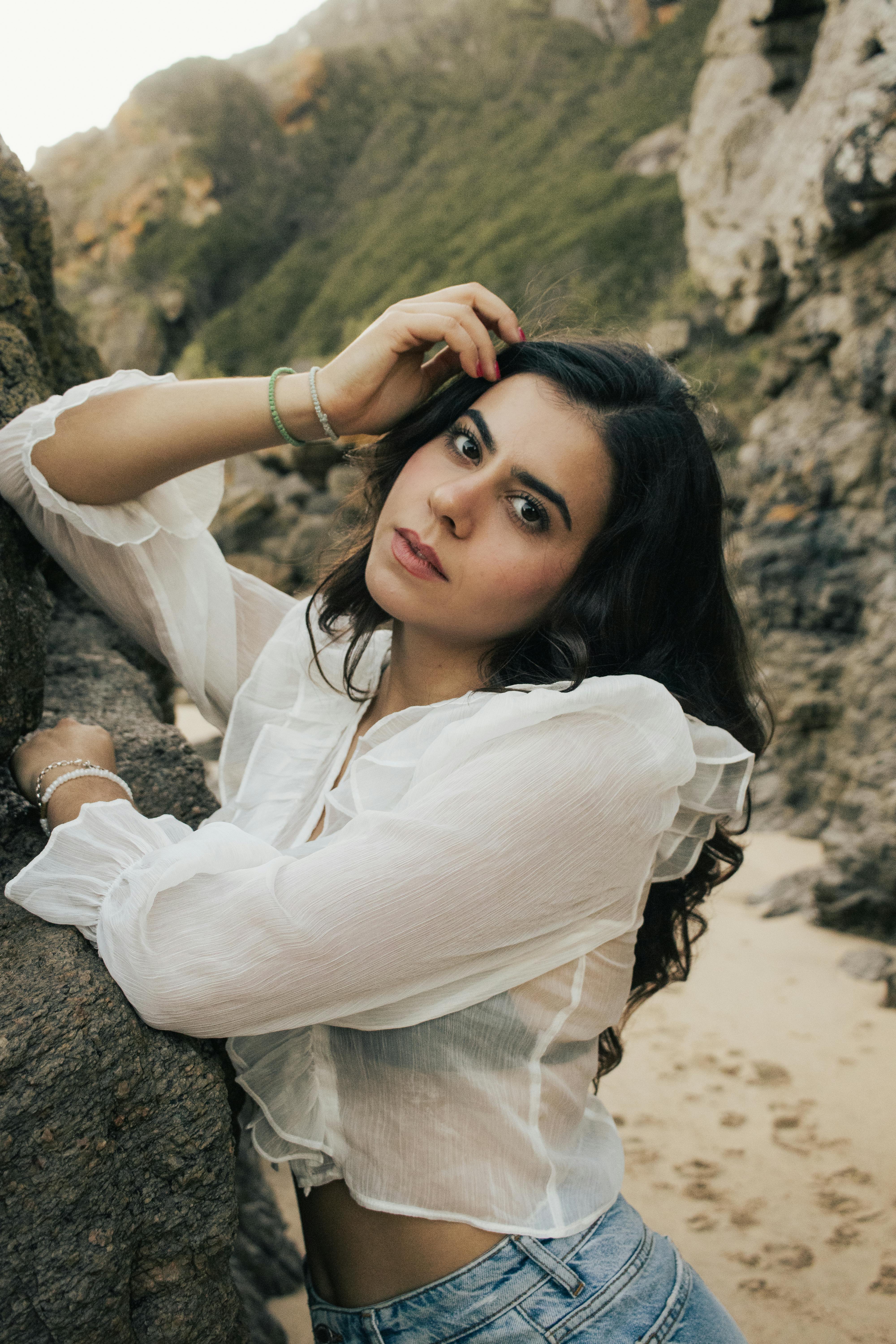 A woman in a white blouse poses by rocks on a scenic beach.