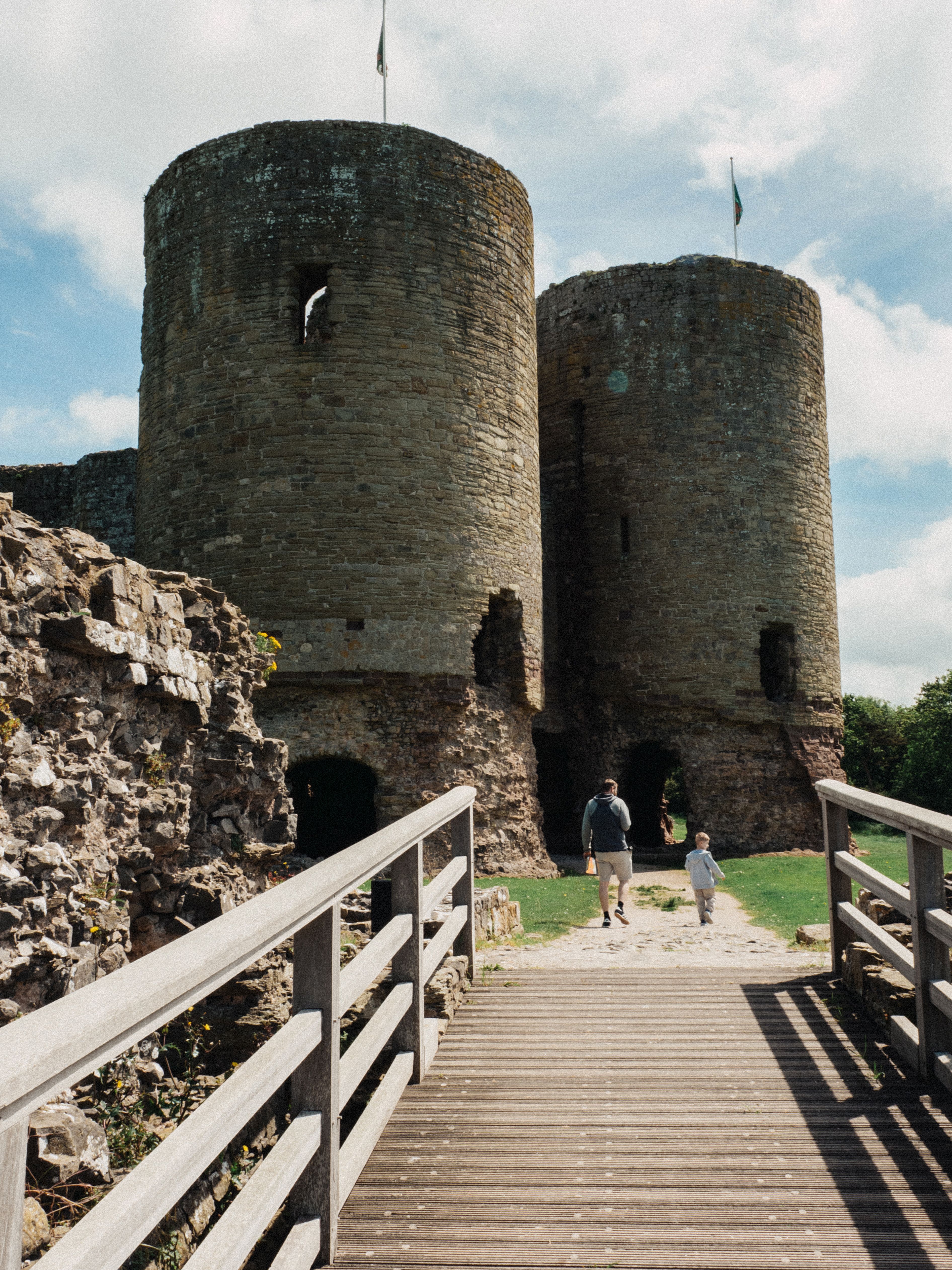 Scenic view of Rhuddlan Castle's twin towers with a wooden bridge and visitors under a sunny sky.