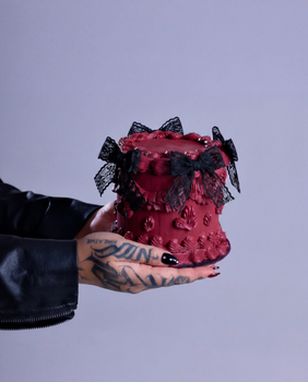 A gothic-themed mini cake, adorned with black lace bows, held by tattooed hands.