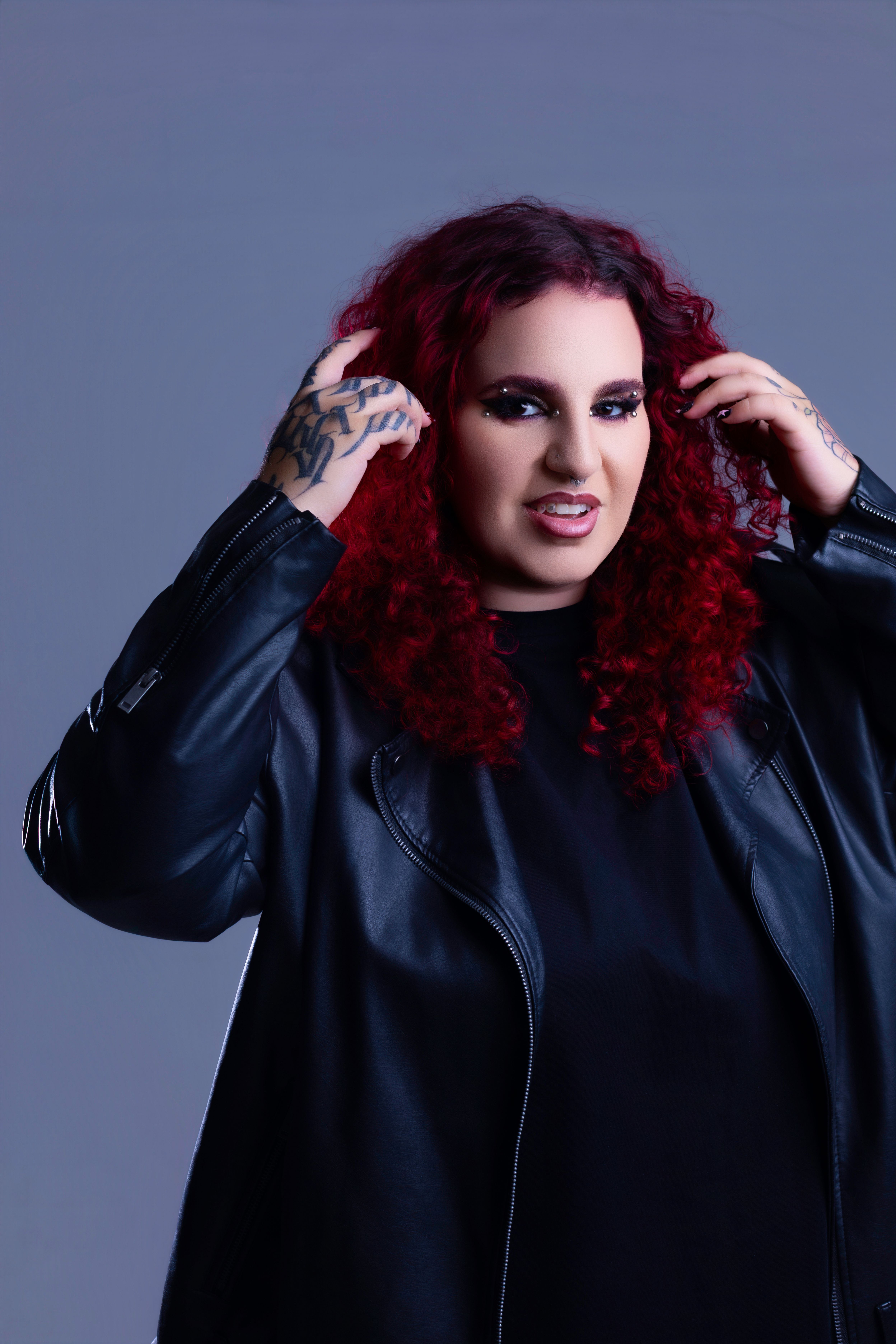 Woman with vibrant red curly hair, wearing leather jacket, tattoos visible.