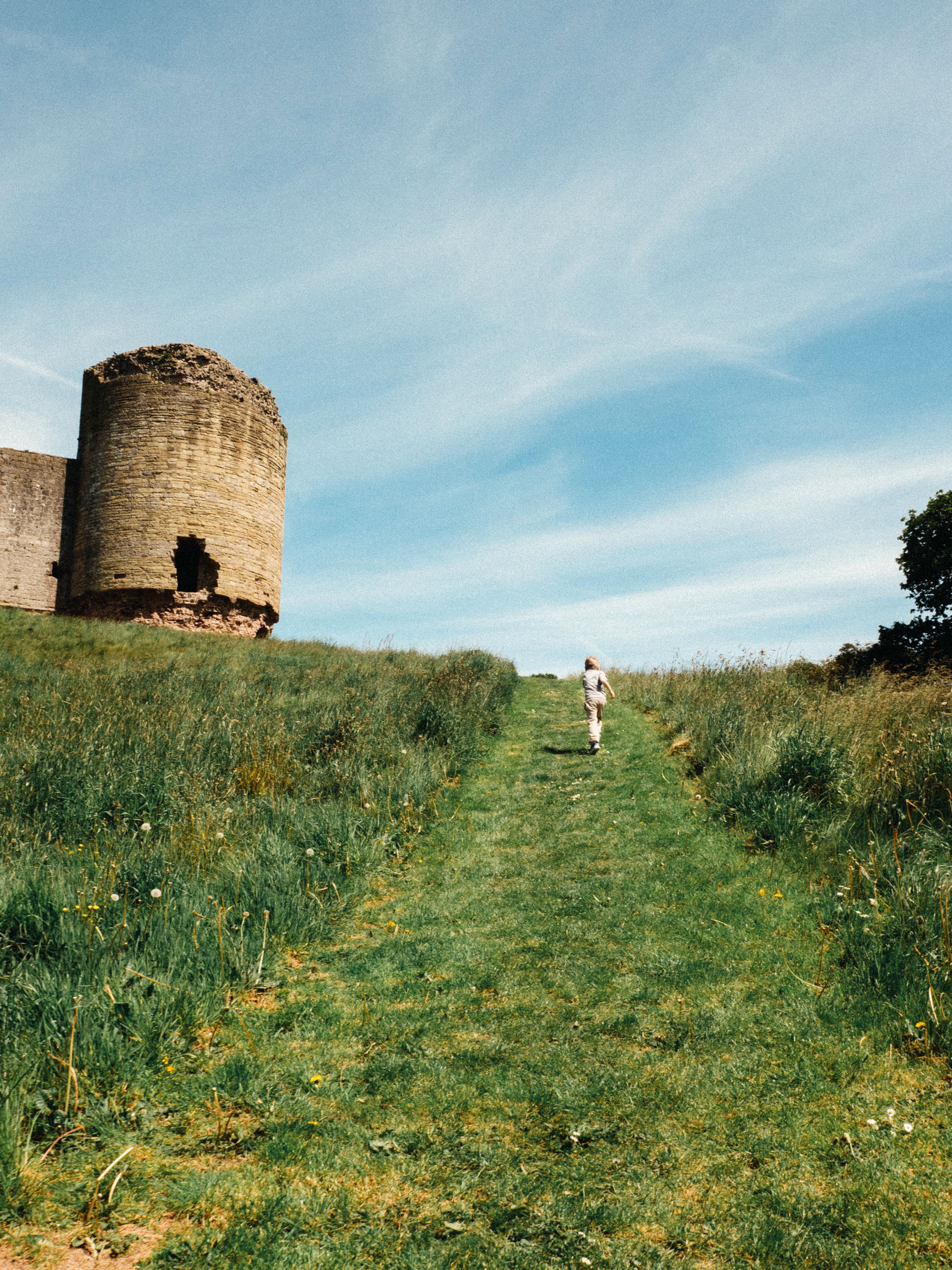 A lone figure walks toward the ruins of Rhuddlan Castle in a grassy field under a blue sky.