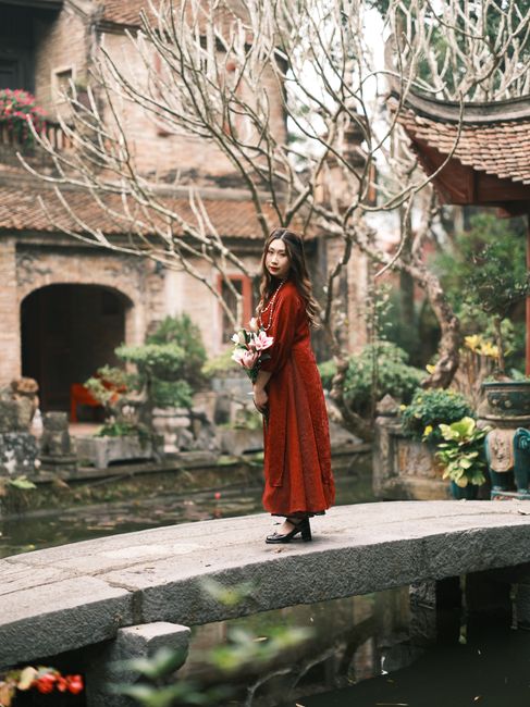 Woman in red dress stands gracefully on bridge in Vietnamese garden.